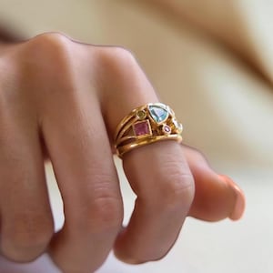 Könnte beinhalten: Ein Goldring mit einem mehrteiligen Steindesign, bestehend aus einem dreieckigen hellblauen Stein, einem quadratischen rosa Stein, einem kleinen runden grünen Stein und einem kleinen runden rosa Stein auf einer polierten Goldschiene.