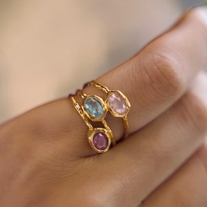 Könnte beinhalten: Ein Set aus drei Goldringen mit einer texturierten, organischen Ringschiene. Jeder Ring ist mit einem einzelnen oval geschliffenen Edelstein in einer Zargenfassung versehen. Die Steine sind hellblau, blassrosa und tief magentafarben.
