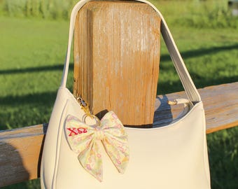 Puede incluir: Un bolso de hombro de color crema con una sola correa y cierre de cremallera superior. En el lateral lleva un lazo de tela decorativo con un estampado amarillo claro y las letras griegas rojas Chi Omega, XΩ, en una anilla de metal dorado.