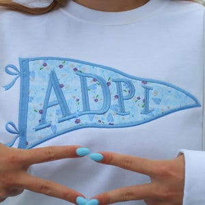 May include: A white crewneck sweatshirt featuring a large, light blue pennant-shaped applique on the chest. The pennant has a light blue background with a small floral pattern and the letters ADPI embroidered in a bold, matching light blue thread.