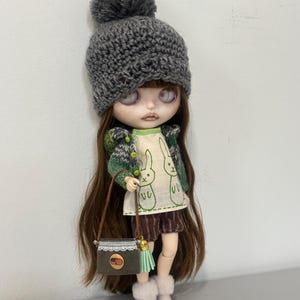 Könnte beinhalten: Eine Puppe mit langem braunem Haar trägt eine graue Strickmütze mit Bommel, eine grün-weiß gemusterte Strickjacke, ein cremefarbenes Oberteil mit grünen Hasenmotiven, braun gestreifte Shorts und helle Stiefel sowie eine kleine graue Cordtasche.
