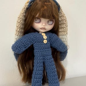 May include: A doll with long brown hair wearing a blue crocheted jumpsuit with three light-colored buttons on the chest and a matching blue crocheted headband with long, beige and white striped crocheted bunny ears extending from the sides.