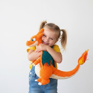 May include: A handmade orange and cream crochet dragon plush toy with blue eyes and a yellow and orange flame tail. The dragon is being held by a smiling child wearing a yellow shirt and blue overalls.