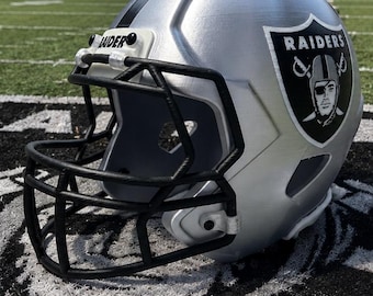 Puede incluir: Un casco de fútbol americano plateado con una rejilla protectora negra y el logotipo de los Raiders en el lateral. El logotipo muestra el texto RAIDERS sobre la ilustración de una persona con un parche en el ojo y un casco. Una franja negra.