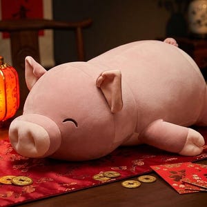 May include: A large, plush pink pig on a red table runner. The pig has a smiling face and is surrounded by oranges, gold coins, and a lit red lantern. The scene evokes a festive, celebratory atmosphere.