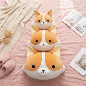 May include: Three plush corgi dog pillows stacked on a pink surface. The pillows are orange and white with black eyes and a black nose. A beige blanket, wheat stalks, a pair of pink slippers, and two open magazines are also in the image.