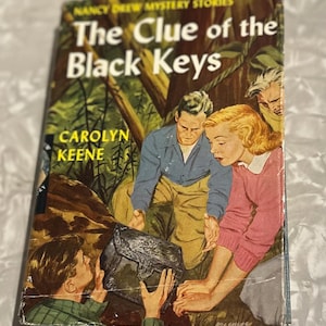 May include: Vintage hardcover book titled "The Clue of the Black Keys" by Carolyn Keene. The cover art depicts a group of people examining a black chest in a forest setting. The book has a worn appearance.