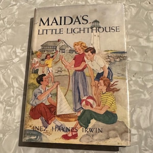 May include: Vintage book cover titled "Maida's Little Lighthouse" by Inez Haynes Irwin. The cover art depicts a coastal scene with children, a lighthouse, and a sailboat. The book has a worn appearance.