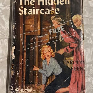 May include: Vintage hardcover book titled "The Hidden Staircase" from the Nancy Drew Mystery Stories series. The cover art features a blonde woman kneeling, pointing, and looking surprised, with two other women in the background. The book has a distressed appearance.