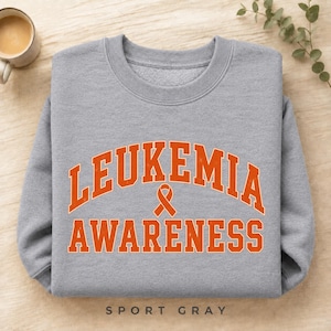 May include: A heather gray crewneck sweatshirt featuring the orange text LEUKEMIA AWARENESS in a collegiate font, arched over an orange awareness ribbon symbol. The design is centered on the chest of the long-sleeved pullover garment.