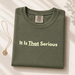 Könnte beinhalten: Ein olivgrünes T-Shirt mit Rundhalsausschnitt und dem hellgelben Aufdruck "It Is That Serious" auf der Brust. Das T-Shirt hat ein Comfort Colors Markenetikett am inneren Halsausschnitt, das die Größe Large angibt, und wird mit einer Goldkette kombiniert.