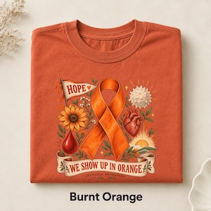 Può includere: Una t-shirt color arancio bruciato con una stampa grafica raffigurante un nastro di sensibilizzazione arancione, un girasole, un cuore, una goccia di sangue e un sole. Il testo recita HOPE e WE SHOW UP IN ORANGE LEUKEMIA AWARENESS con motivi floreali.