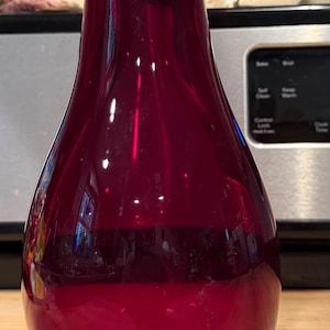 Puede incluir: Un jarrón de cristal rojo rubí translúcido, alto, con un cuerpo liso y bulboso que se estrecha hacia un cuello delgado. La base es gruesa y transparente, proporcionando una base estable para el recipiente. El cristal tiene un acabado pulido.
