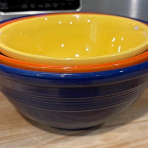 May include: A set of three nested ceramic bowls in vibrant colors. The largest bowl is a deep blue, followed by an orange bowl, and a bright yellow bowl nested inside. The bowls have a smooth, glossy finish and are stacked on top of each other.