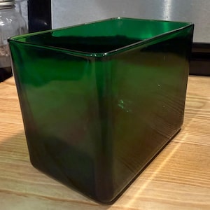 May include: A rectangular glass vase in a deep emerald green color. The vessel features smooth, straight sides with rounded corners and an open top, showcasing a translucent quality that allows light to pass through the thick, solid glass walls.