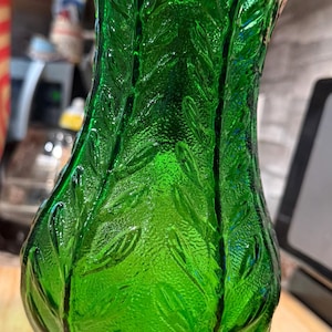 Peut inclure: Un vase en verre vert avec un motif de feuilles texturées. Le vase a une base bombée et un dessus évasé. Le verre est d'un vert vif et le motif de feuilles est en relief.