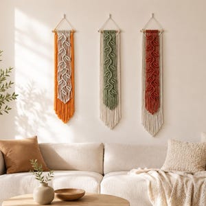 May include: Three vertical macrame wall hangings suspended from wooden dowels. Each piece features a central leaf-like pattern in orange, sage green, and terracotta, layered over cream-colored cord with a long, fringed bottom edge.