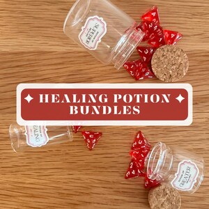 Peut inclure: Trois flacons en verre transparent avec bouchons en liège et étiquettes Potion of Superior Healing, Potion of Healing et Potion of Greater Healing. Des dés polyédriques rouges translucides sont éparpillés. Une bannière indique HEALING POTION BUNDLES.