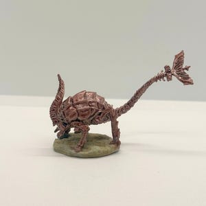 May include: A detailed, reddish-brown miniature sculpture of a creature with a segmented body, long neck, and a fin-like structure at the end of its tail. The figure is posed on a small, textured base, suggesting a prehistoric or fantasy theme.