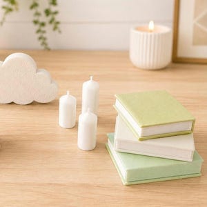 May include: A stack of three small rectangular books with light green and white covers, accompanied by three white pillar candles and a white cloud-shaped object. A single lit white candle in a ribbed holder sits in the background.