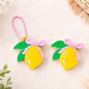 May include: Two lemon-shaped charms featuring a bright yellow lemon, green leaves, and a light pink bow. One charm includes a pink ball chain attachment. The design is flat with defined outlines, showcasing a vibrant fruit motif with a decorative ribbon.