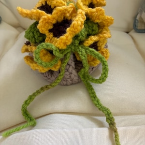 May include: A crocheted bouquet of sunflowers. The sunflowers have yellow petals with dark brown centers and green leaves. The bouquet is tied with a green yarn bow and sits in a gray crocheted pot.