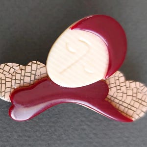 Peut inclure: Broche vintage au design stylisé. Elle présente un visage crème avec des détails gravés, une coiffe bordeaux brillante et une base incurvée bordeaux. La broche comprend deux éléments beiges à motifs de mosaïque.