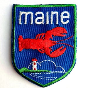 May include: A shield-shaped embroidered patch with a blue background and a green border. It features the white text "maine" at the top, a large red lobster in the center, and a small white and red lighthouse on a green hill above blue water.
