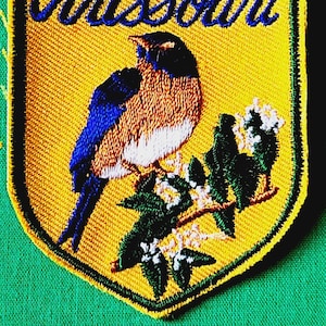 May include: A shield-shaped embroidered patch with a yellow background and a dark green border. The word Missouri is stitched in blue script at the top. A blue and orange bird is perched on a branch with green leaves and white flowers.