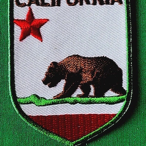 May include: A shield-shaped embroidered patch featuring the word CALIFORNIA in black text at the top. The design includes a red star on a white background above a brown bear walking on a green patch of grass, with a solid red horizontal stripe below.