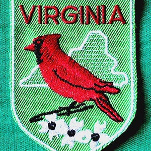 May include: A shield-shaped embroidered patch with a light green background. It features the red text VIRGINIA at the top, a red cardinal bird perched on a branch in the center, and white dogwood flowers with black centers at the bottom.