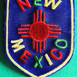 May include: A shield-shaped embroidered patch with a blue background and a yellow border. It features a red Zia sun symbol in the center. The word NEW is stitched in red and yellow at the top, with MEXICO in yellow and green below.