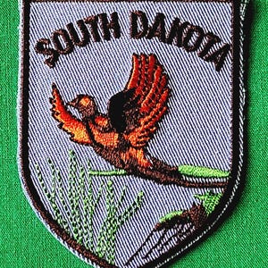 May include: A shield-shaped embroidered patch featuring a light blue background with the text SOUTH DAKOTA arched at the top. The design depicts a pheasant in flight with orange, brown, and black plumage above green grass and foliage.