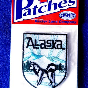 May include: An iron-on embroidered patch in a shield shape featuring the word Alaska in black text above a graphic of a black and white husky dog standing in a snowy landscape with mountains in the background, packaged on a red and white card.
