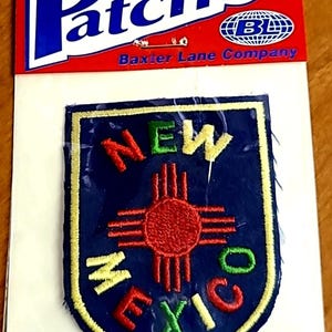 May include: Iron-on patch featuring a shield design with the words "NEW MEXICO" in colorful embroidery. The patch has a blue background with a red sun symbol in the center and is packaged on a red and white backing.