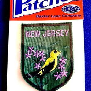 May include: Iron-on patch featuring a shield design with the text "NEW JERSEY" in pink embroidery. The patch showcases a yellow and black bird with purple flowers on a green background. The patch is from the Baxter Lane Company.