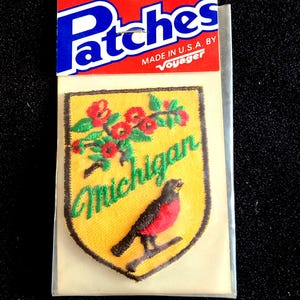 May include: Iron-on patch featuring a yellow shield with a red bird and floral design, with the word "Michigan" in green script. The patch is packaged with a red and blue header that reads "Patches" and "Iron-On". Made in the U.S.A. by Voyager.
