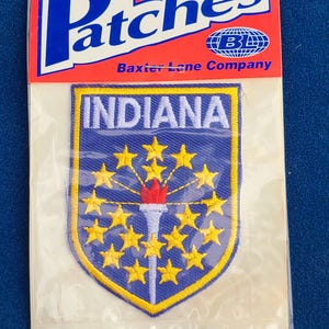 May include: An iron-on patch featuring the state of Indiana. The patch has a blue background with a yellow border and stars. The word "INDIANA" is across the top. The patch is in a clear plastic package with a red and white header that says "Patches".