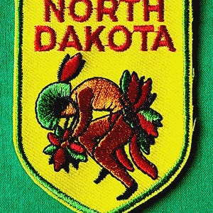 May include: A shield-shaped embroidered patch with a bright yellow background and a green border. The text NORTH DAKOTA is stitched in red at the top. Below the text is a multi-colored embroidered figure in a dynamic, bent-over pose.