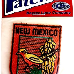 May include: An iron-on patch featuring the state of New Mexico. The patch is orange with a yellow bird and a cluster of yellow and green plants. The text "NEW MEXICO" is at the top. The packaging reads "Patches" and "Baxter Lane Company."