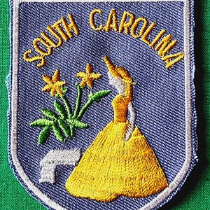 May include: A shield-shaped embroidered patch with a light blue background and a white border. It features the yellow text SOUTH CAROLINA, a yellow-dressed figure, green flowers, and a small white L-shaped element stitched onto the fabric.