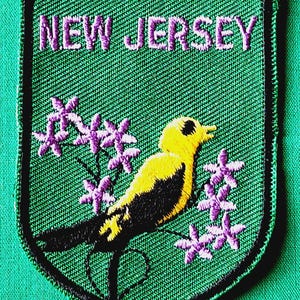 May include: A shield-shaped embroidered patch with a green background and a black border. The text NEW JERSEY is stitched in purple at the top. A yellow and black bird is perched on a branch surrounded by small purple flowers on both sides.