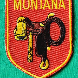 May include: A shield-shaped embroidered patch with a red background and yellow border. The word MONTANA is stitched in yellow at the top. Below the text is a black and yellow embroidered graphic of a western saddle with a coiled lasso attached.
