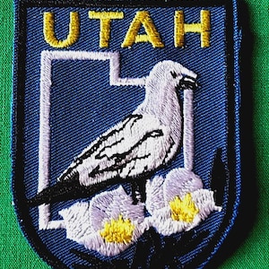 May include: An embroidered patch featuring a blue background with the word UTAH in gold letters at the top. The design includes a white outline of the state of Utah, a white seagull, and two white flowers with yellow centers at the bottom.