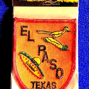 May include: An orange iron-on cloth emblem patch featuring the text EL PASO TEXAS in black embroidery. The patch includes a yellow roadrunner bird and a yellow sombrero with a green brim. The packaging is labeled IRON ON CLOTH EMBLEM.