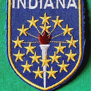 May include: A shield-shaped embroidered patch featuring a blue background with the word INDIANA in white letters at the top. A central white torch with a red flame is surrounded by nineteen gold embroidered stars arranged in a circular pattern.