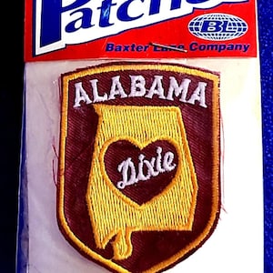 May include: An iron-on patch featuring the state of Alabama in a shield shape. The patch has a maroon border with the word "ALABAMA" in white. Inside the state outline is a heart with the word "Dixie". The patch is in a clear plastic package with the word "Patches" on top.