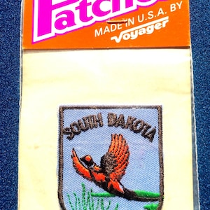 May include: Iron-on patch featuring a South Dakota design with a flying bird and the words "South Dakota". The patch is on a blue background with green grass. The patch is packaged on a card that says "Patches" and "Made in U.S.A. by Voyager".