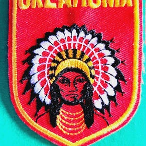 May include: A shield-shaped embroidered patch with a red background and yellow border. The text OKLAHOMA is stitched in yellow at the top. Below, a figure wears a large, detailed white, black, and yellow feathered headdress and yellow necklace.