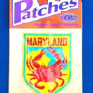 May include: An iron-on embroidered patch in a shield shape featuring a red crab on a yellow background with the word MARYLAND in red text. The packaging is labeled Patches, Baxter Lane Company, and includes an iron-on icon at the top.